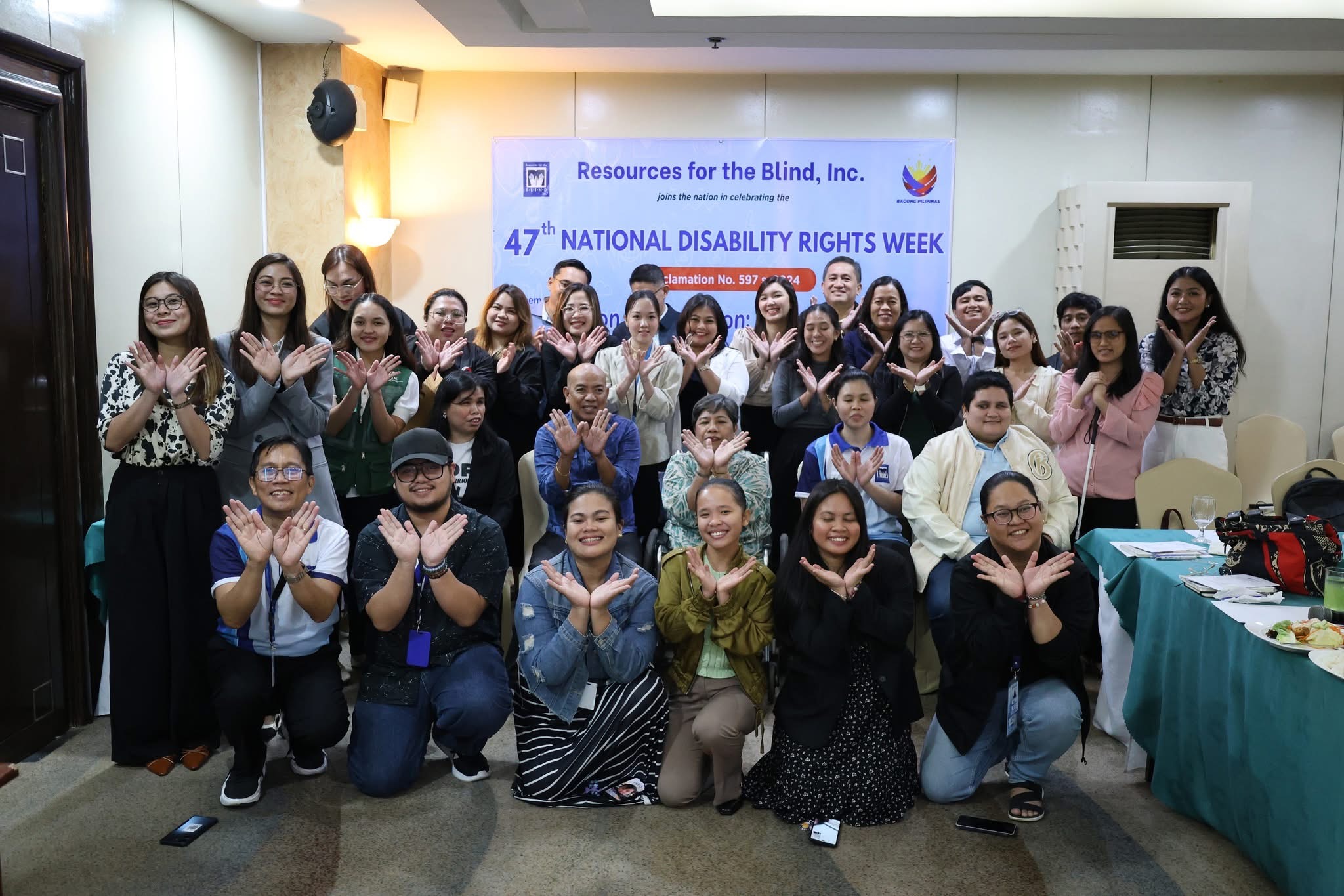 A photo captures a big group of people making hand gestures that form the logo of RBI. Behind them, a banner announces that "Resources for the Blind, Inc. joins the nation in celebrating the 47th National Disability Rights Week."